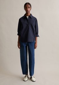 Marineblaues Baumwollhemd mit geknöpftem Kragen und Brusttasche, kombiniert mit blauen Denimhosen und weißen Slip-On-Schuhen.