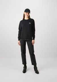 Felpa nera con colletto a girocollo e polsini a coste, abbinata a jeans scuri e stivali alla caviglia. Un cappellino nero completa il look informale.