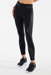 Leggings atletici neri con texture opaca e lucida a contrasto, vita alta e design a pannelli curvi. Abbinati a scarpe da ginnastica nere.