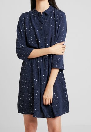 Kobieta ubrana w granatową sukienkę z kołnierzykiem, zapinaną na guziki, w białe grochy, z rękawami do połowy przedramion, stojąca z założonymi rękami.