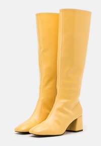 Bottes jaunes montantes en matériau brillant, avec un bout carré et un block heel épais. Texture lisse avec des détails de couture minimalistes.
