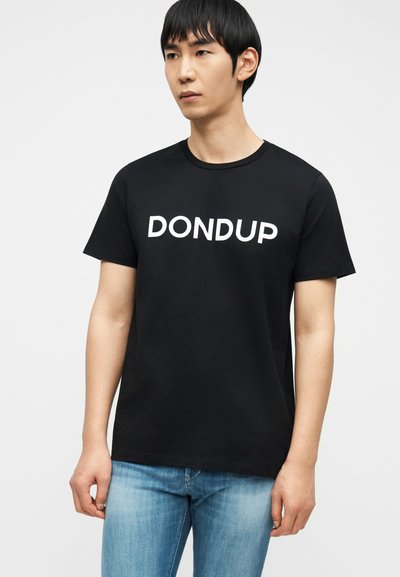 Άνδρας με μαύρο T-shirt με λευκό κείμενο "DONDUP" και ανοιχτόχρωμο μπλε τζιν, στέκεται μπροστά σε απλό ανοιχτόφωτο φόντο, κοιτάζοντας ελαφρώς προς τα δεξιά.