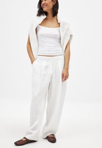 Weiße Leinen-Hose mit weitem Bein, kombiniert mit einem figurbetonten weißen Tanktop und einem leichten, offenen Strickschal. Braune Ballerinas runden das Outfit ab.
