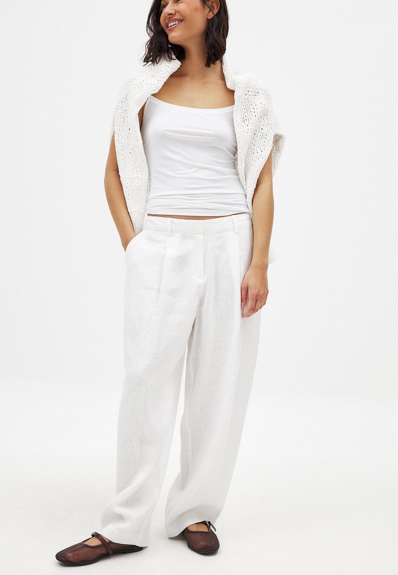 Weiße Leinen-Hose mit weitem Bein, kombiniert mit einem figurbetonten weißen Tanktop und einem leichten, offenen Strickschal. Braune Ballerinas runden das Outfit ab.