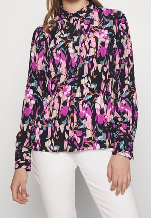 Woman wearing a long-sleeve button-up blouse with a colorful abstract pattern in pink, purple, blue, and beige, paired with white pants.