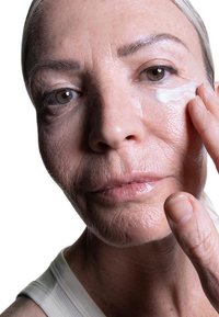Close-up of a person applying white cream to their cheek. Visible fine lines and a smooth texture on the skin. Light background.