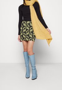 Haut noir à manches longues, écharpe jaune texturée, jupe verte imprimée léopard jaune, bottes bleu marine montantes avec une finition brillante et un bout pointu.