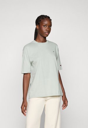 Donna con i capelli intrecciati che indossa una t-shirt oversize grigio chiaro e pantaloni crema, in piedi davanti a uno sfondo chiaro e uniforme.