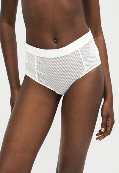 Γυναίκα που φοράει λευκά εσώρουχα με υψηλή μέση και λεπτές κάθετες ρίγες, στέκεται μπροστά σε ένα απλό φωτεινό φόντο.