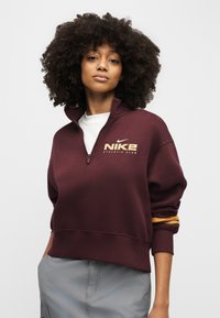 Noor naine lokkis juustega, kandes burgundia värvi Nike Athletic Club poollukuga pusat ja heledat halli pükse, seistes ühevärvilise tausta ees.