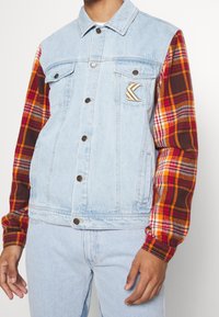 Denim jacket with light blue body, plaid fabric sleeves in red, orange, and brown, featuring two chest pockets and branded detail.