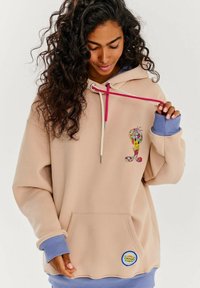Béžová mikina s ružovou a krémovou šnúrkou, jazdí farebným kresleným dizajnom na hrudi a modrými akcentmi na rukávoch a leme.