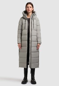 Lang, lys grå pufferjakke med hætte, quiltet design, frontlynlås og snøre detaljer. Bæres over en mørk top og sorte støvler.