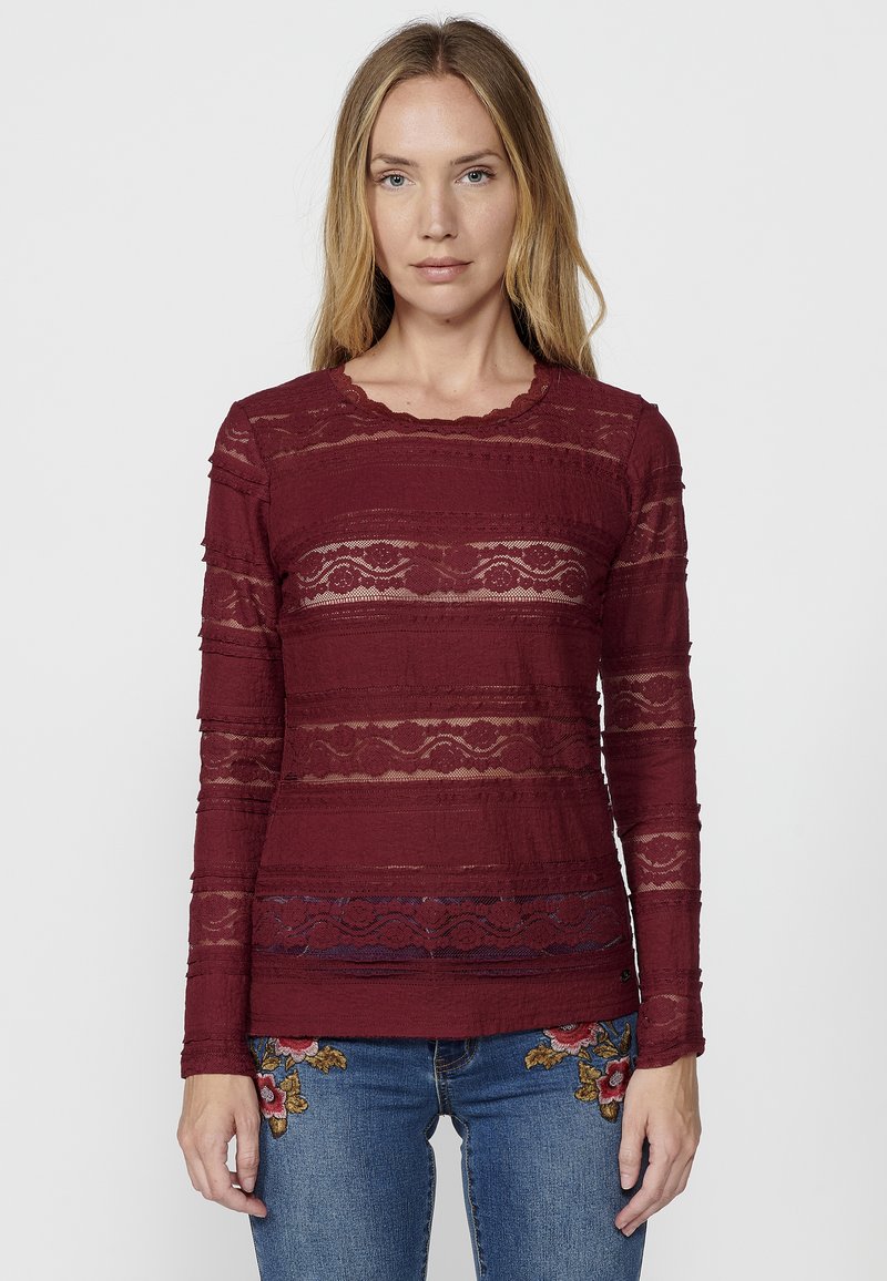 Top a maniche lunghe color bordeaux con strisce di pizzo, scollo a onde, tessuto texturizzato, abbinato a jeans ricamati con dettagli floreali.