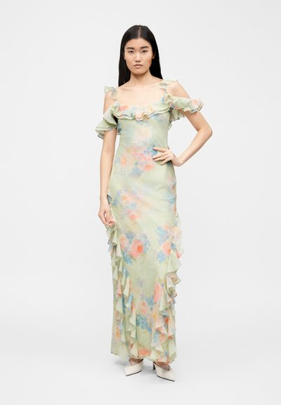 Γυναίκα στέκεται με το χέρι στη μέση, φορώντας παστέλ πράσινο φόρεμα με λουλούδια και ακάλυπτους ώμους, με βολάν και λευκά μυτερά τακούνια, σε λευκό φόντο.