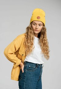 Żółta czapka beanie z logo, dżinsowa kurtka i biała koszulka. Modelka ma długie, falujące włosy i nosi spodnie dżinsowe z wysokim stanem w kolorze niebieskim.