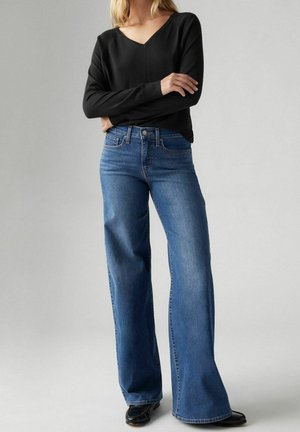 Personne portant un haut noir à manches longues avec un col en V et un jean bleu taille haute à jambes larges, debout les bras croisés devant un fond uni.