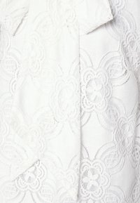 Vit blus med intrikata spetsdesign som har blommönster och upphöjd sömnad. Mjuk, texturerad tyg med en svävande drapering.