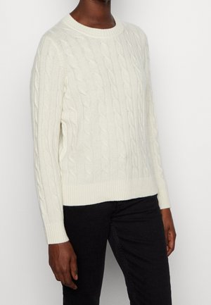 Personne portant un pull blanc à torsades à manches longues et un pantalon noir sur un fond clair uni.