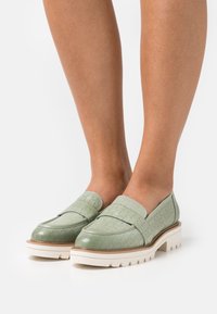 Mocassins en cuir tissé vert clair avec semelles blanches épaisses portés pieds nus sur un fond blanc.