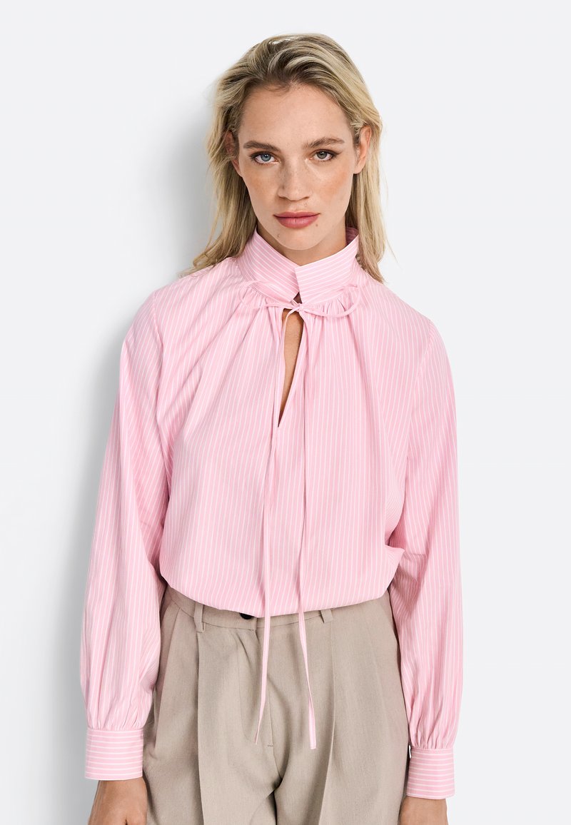 Blusa lunga a maniche di colore rosa chiaro con righe bianche verticali, colletto alto, apertura centrale a forma di chiave e dettaglio annodato al collo. Tessuto morbido e vestibilità ampia.