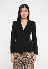 Blazer noir avec col à revers et détails en métal doré à la taille, associé à un pantalon à imprimé léopard, coupe structurée et texture lisse.