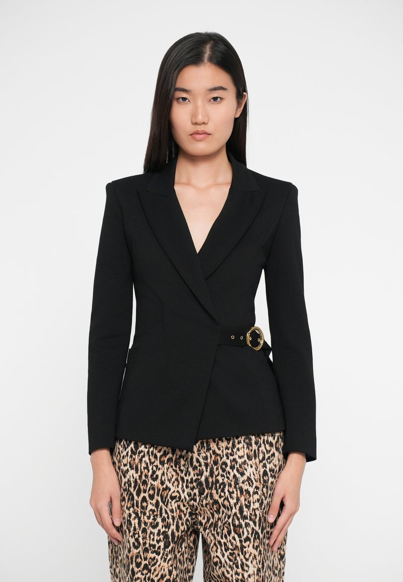 Blazer noir avec col à revers et détails en métal doré à la taille, associé à un pantalon à imprimé léopard, coupe structurée et texture lisse.
