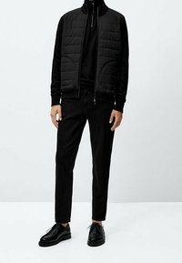 Homme portant une veste matelassée noire par-dessus un pull à fermeture éclair noir, un pantalon slim noir et des chaussures en cuir noires, debout devant un fond uni.