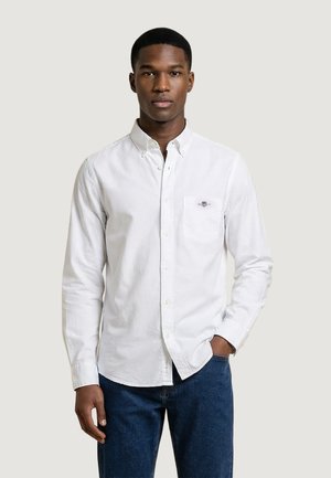 Mężczyzna ubrany w białą koszulę z guzikiem, kieszenią i małym logo, w połączeniu z niebieskimi dżinsami, stojący na tle jednolitego jasnego tła.