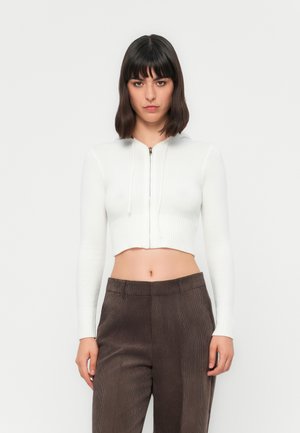 Vrouw met schouderlang donker haar, gekleed in een witte cropped ritstrui en high-waisted bruine corduroy broeken, staand tegen een effen achtergrond.