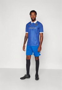 Uomo in piedi con divisa da calcio blu, scarpe da calcio nere e calzini neri, sfondo neutro, rivolto in avanti con espressione neutra.