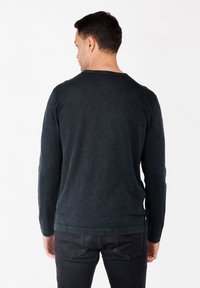 Hombre de espaldas vistiendo una camiseta de manga larga oscura y jeans negros desgastados, frente a un fondo blanco liso.