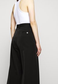 Los pantalones de pierna ancha negros de tiro alto presentan una textura suave, un bolsillo trasero con detalle de botón y acentos de pliegue.