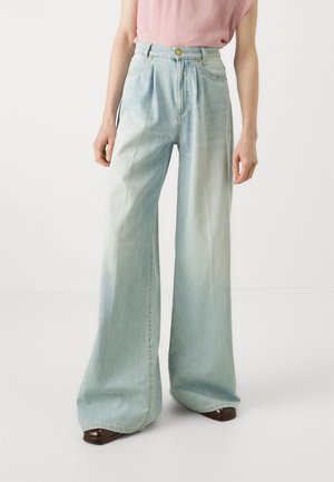 Personne portant un jean taille haute bleu clair à jambes larges, associé à un haut rose à manches courtes et des chaussures marron foncé.