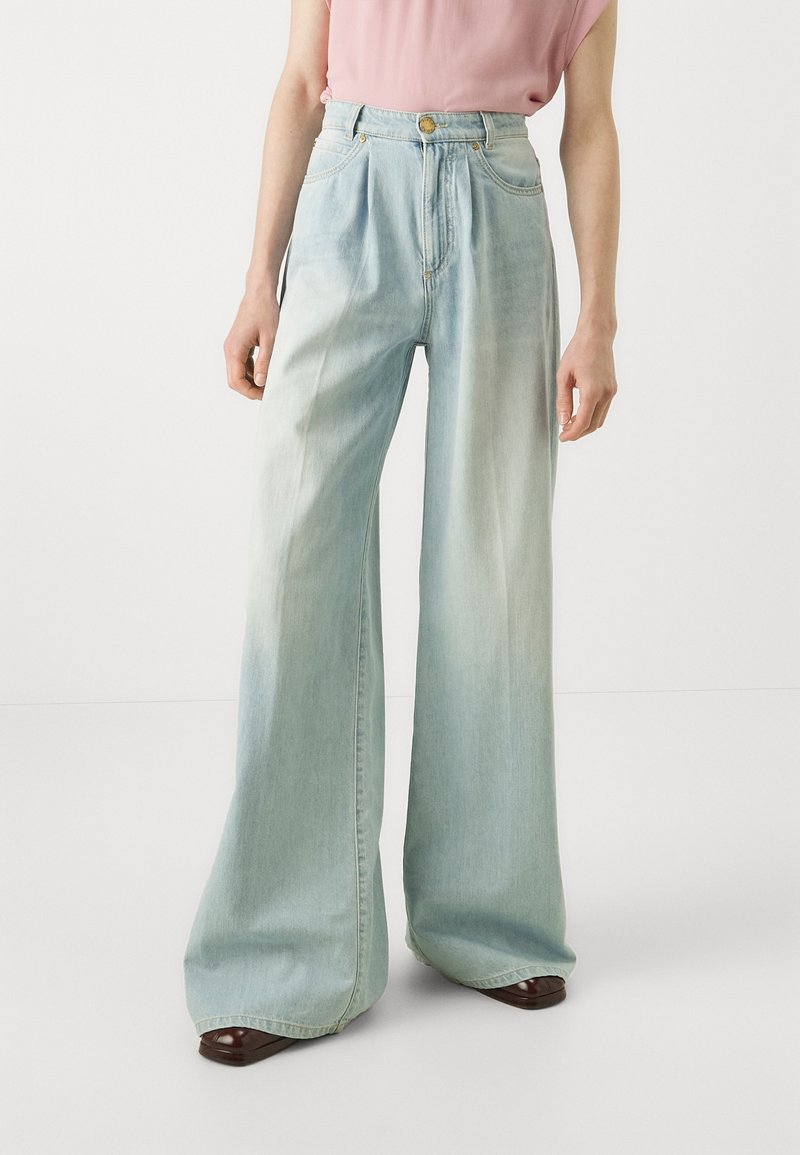 Personne portant un jean taille haute bleu clair à jambes larges, associé à un haut rose à manches courtes et des chaussures marron foncé.