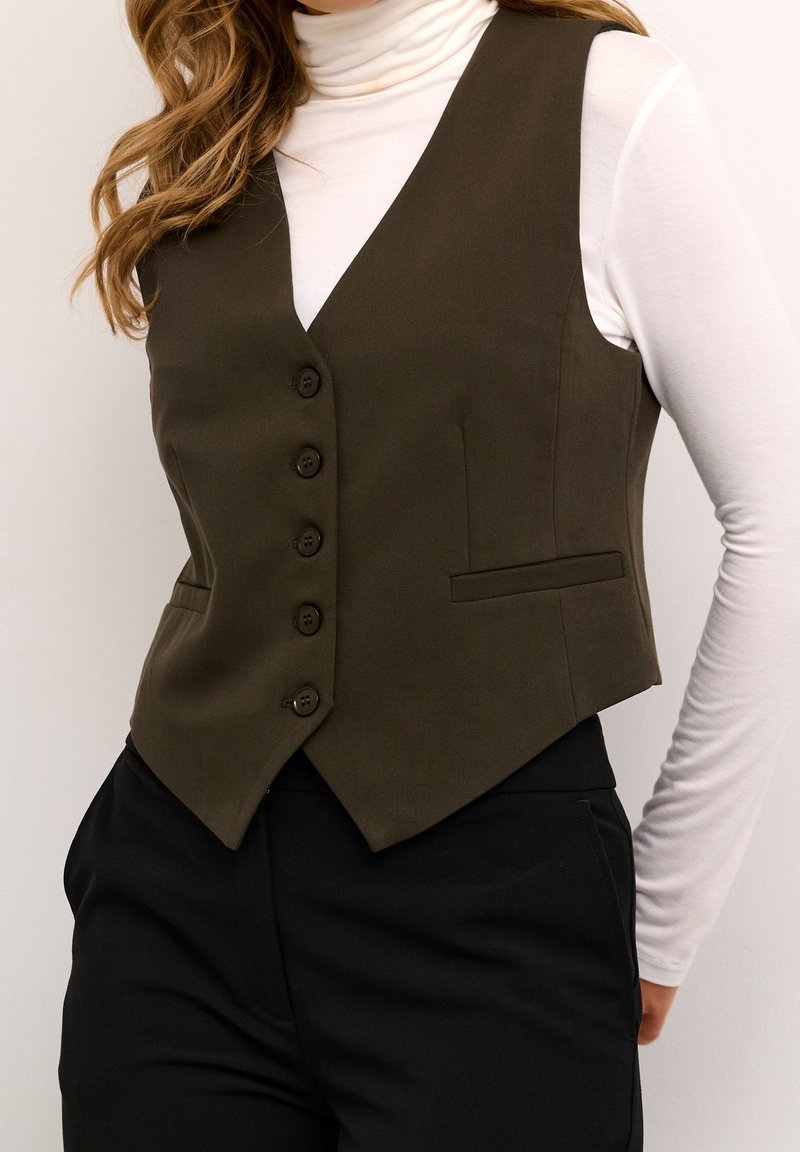 Femme portant un gilet boutonné olive foncé sur un col roulé blanc et un pantalon noir, avec des cheveux ondulés châtain clair visibles.