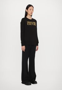 Melna džemperis ar zelta "VERSACE JEANS COUTURE" uzrakstu, apaļš kakla izgriezums, ribotām piedurknēm, kopā ar platām melnām biksēm un masīvām melnām kurpēm.