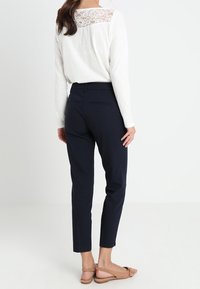 Vit långärmad topp med spetsdetalj på ryggen, i kombination med marinblå smala byxor och beige platta skor. Enkelt, elegant mönster med mjuka texturer.