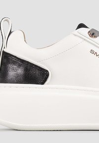 Sneaker bianco con accento in pelle nera, superiore testurizzato, punta tonda e suola piatta in gomma. Presenta un'etichetta di presa e cuciture decorative.