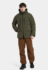 Doudoune vert olive avec fermeture éclair sur le devant, deux poches poitrine et une capuche ; associée à un pantalon brown ample et des chaussures noires.