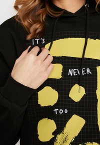 Sudadera negra con un patrón de cuadrícula, que presenta pinceladas amarillas de gran tamaño y el texto blanco "NUNCA ES DEMASIADO." El texto está centrado.