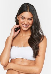 Valge ilma õlakuputeta bandeau-top, millel on pitsäär ja tekstuuriga kangas, featuring lillemuster ja kehaümberügav disain. Modell poseerib käsi kaelal.