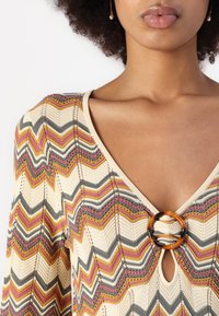 Top en maille avec décolleté en V, motif en zigzag dans des teintes beige, gris, rose et jaune, accentué par un anneau circulaire en écaille.