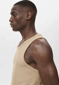 Beige tanktop med V-hals, ribbet tekstur og brede ærmegab, præsenteret på en model mod en neutral baggrund.