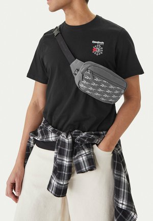 Hombre con camiseta negra de Reebok, bolso bandolera gris con estampado, pantalones blancos y camisa a cuadros en blanco y negro atada a la cintura.