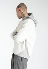 Sweatshirt med hvidt krop og grå hætte. Kasseformet design, blød tekstur og løs pasform. Et par grå bukser ses nedenunder.