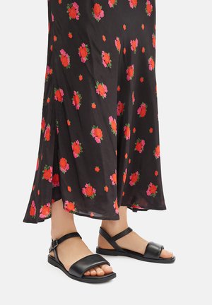 Černé maxi šaty s květinovým motivem v červené a růžové barvě, doplněné černými sandály na ploché podrážce se páskem přes chodidlo a kolem kotníku.