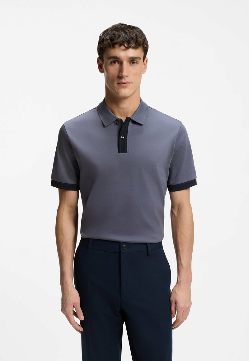 Homme debout face à l'avant, portant un polo gris à manches courtes avec des bordures noires et un pantalon foncé, sur un fond blanc uni.