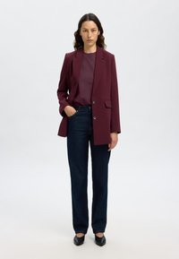 Blazer su misura bordeaux, abbinato a un top viola intenso e jeans dritti in denim scuro. Scarpe nere completano l'outfit.