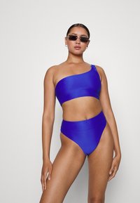 Top de bikini bandeau assimétrico e parte de baixo de bikini alta em tecido texturizado azul brilhante. Inclui um detalhe de fecho no ombro.